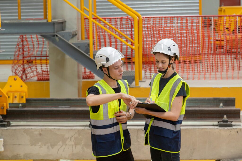 Two construction site workers wearing high-visibility vests and safety helmets reviewing notes on a clipboard in an industrial environment with safety barriers and metal railings.