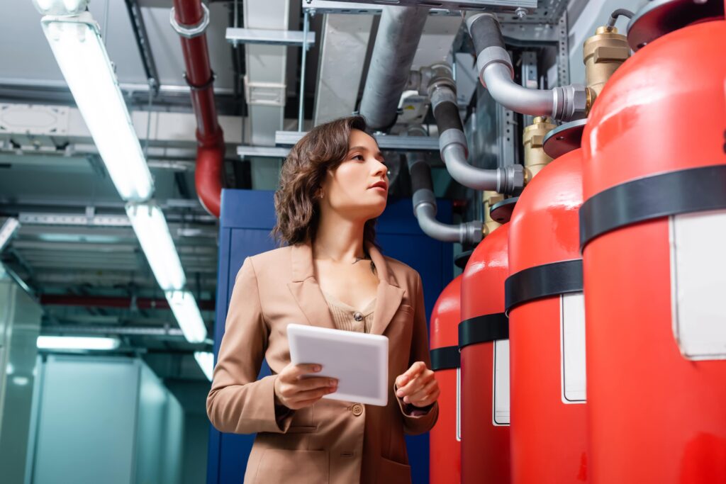 Person holding a tablet inspecting red fire suppression tanks and pipework in a commercial building plant room.